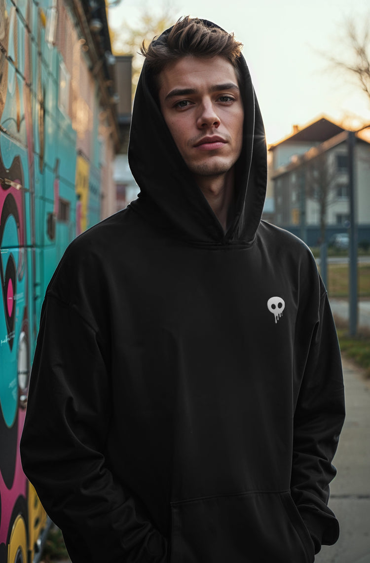 young blonde man wearing a black hoodie with the popsikulls skull logo on a urban environment next to a colorful graffiti wall